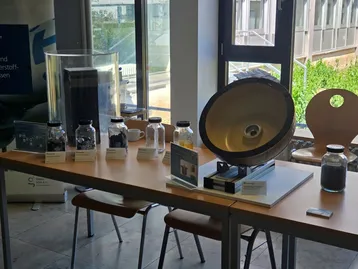 Auf dem Tisch stehen mehrere Gläser mit verschiedenen Proben, darunter ein großer Trichter und ein geschwungenes Metallobjekt, das wie eine Lautsprecheranordnung aussieht. Es scheint sich um eine Ausstellung zu handeln, die sich mit Umwelt- oder Materialwissenschaften beschäftigt.