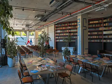 Ein moderner Veranstaltungsraum mit langen Tischen, die mit Prospekten oder Broschüren bedeckt sind. Rote und graue Stühle sind um die Tische gruppiert. Im Hintergrund befinden sich Bücherregale und große Fenster, die viel Tageslicht hereinlassen. Pflanzen sind verstreut im Raum.