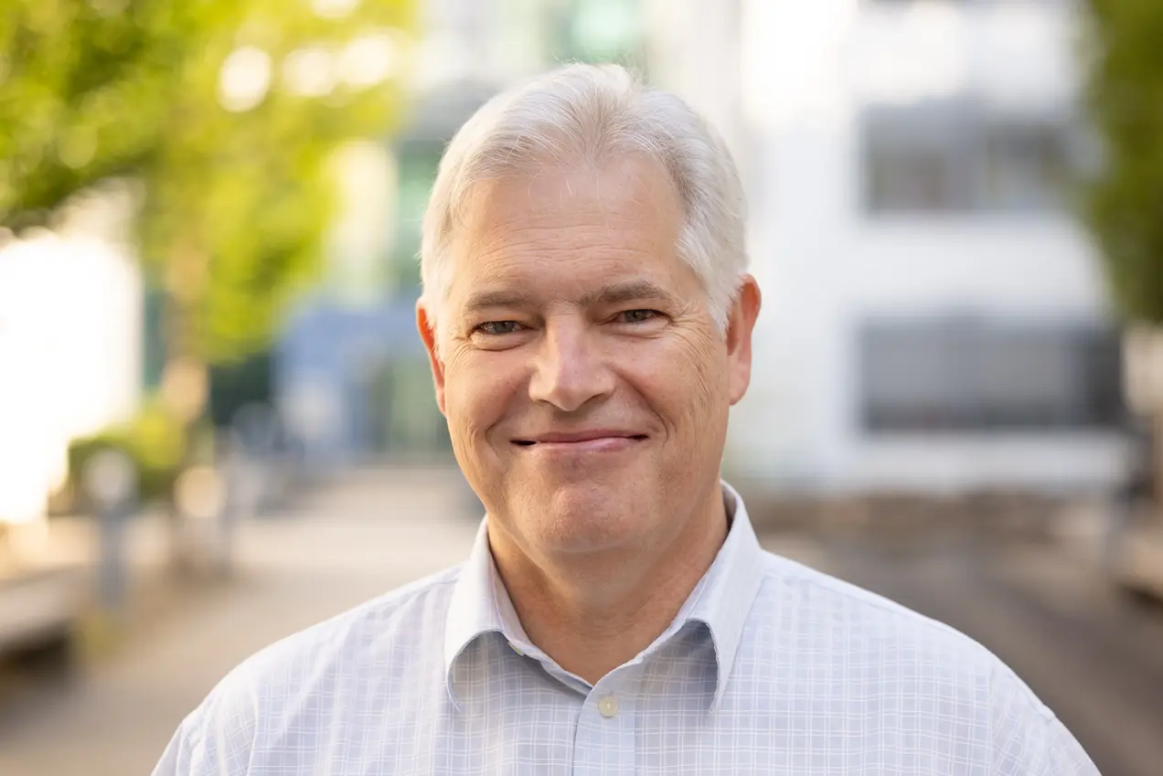 Ein lächelnder Mann mit grauen Haaren und einem hellen Hemd steht in einer urbanen Umgebung. Der Hintergrund enthält unscharfe Gebäude und Bäume. Das Bild vermittelt eine freundliche und offene Atmosphäre.