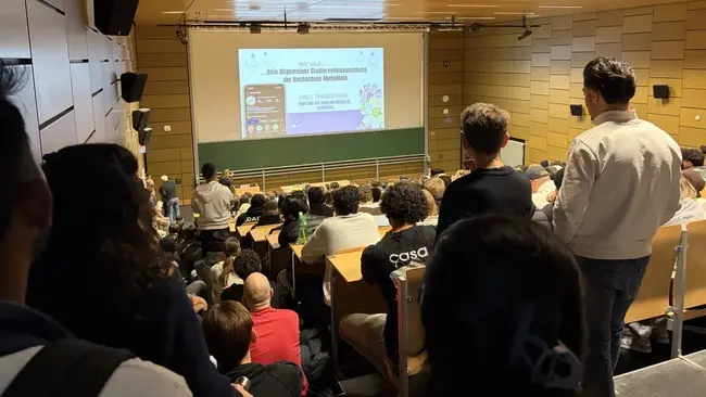 Einblick in einen vollbesetzten Hörsaal mit vielen Menschen, die auf eine Präsentation auf einer Leinwand blicken. Auf der Leinwand sind Informationen zu einer Veranstaltung oder einem Vortrag zu sehen. Die Atmosphäre ist erwartungsvoll und konzentriert.