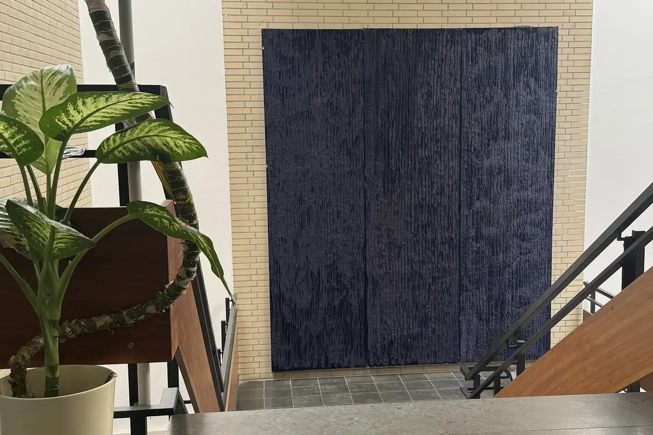 Im Bild ist eine grüne Pflanze in einem weißen Topf zu sehen, die auf einem Tisch steht. Im Hintergrund befindet sich eine blaue Wandverkleidung aus Stoff und eine Treppe führt nach unten. Der Raum ist hell und modern gestaltet.