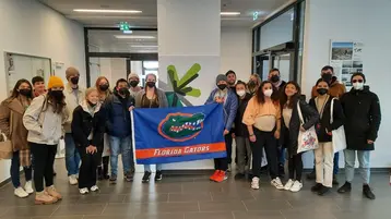 Eine Gruppe von etwa 20 Personen steht in einem Innenraum und lächelt in die Kamera. Einige tragen Schutzmasken. In der Mitte hält jemand eine Flagge der Florida Gators. Der Hintergrund zeigt helle Wände und offene Türen.