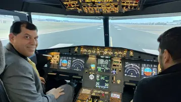 Zwei Männer sitzen in einem Flugzeug-Cockpit. Der Mann auf der linken Seite, in Anzug und Krawatte, lächelt in die Kamera. Der Cockpitbildschirm zeigt Instrumente und Anzeigen für die Fluginformationen. Draußen ist eine Landebahn sichtbar.