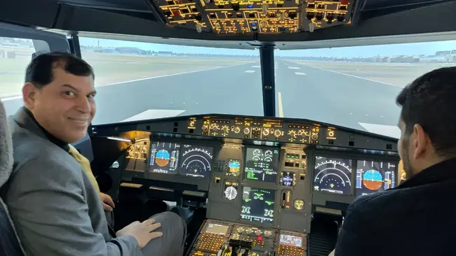 Zwei Männer sitzen in einem Flugzeug-Cockpit. Der Mann auf der linken Seite, in Anzug und Krawatte, lächelt in die Kamera. Der Cockpitbildschirm zeigt Instrumente und Anzeigen für die Fluginformationen. Draußen ist eine Landebahn sichtbar.