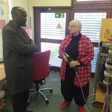 Eine Frau mit Brille und roter Jacke steht an einem Arbeitsplatz und hält ein Gerät in der Hand. Sie spricht mit einem Mann in einem langen, dunklen Mantel. Im Hintergrund ist ein Fenster mit Blättern, ein Sessel und einige Arbeitsgeräte sichtbar.