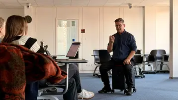 Ein Mann sitzt auf einem Stuhl und spricht zu einer Gruppe von Personen, die ihm gegenüber sitzen. Er gestikuliert mit der Hand, während er Informationen oder Anweisungen gibt, möglicherweise in einem Unterrichts- oder Besprechungskontext.