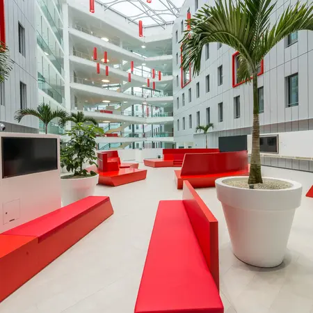 Ein moderner Innenhof mit hellen Wänden und großen Fenstern. Rote Möbel und große Pflanzen sind im Raum verteilt. In der Mitte stehen Sitzbänke in verschiedenen Formen, während ein TV-Gerät an einer Wand platziert ist. Der gesamte Raum wirkt freundlich und einladend.