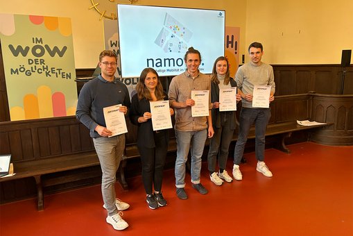 Die Teammitglieder von namowo stehen vor dem Präsentationsbildschirm und halten die Zertifikate in den Händen