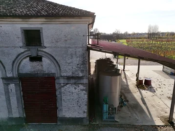 The picture shows an old winery building with a red wooden door and a covered area. Large silver tanks are visible in the foreground. In the background, grapevines and a wooded landscape can be seen under a clear sky.