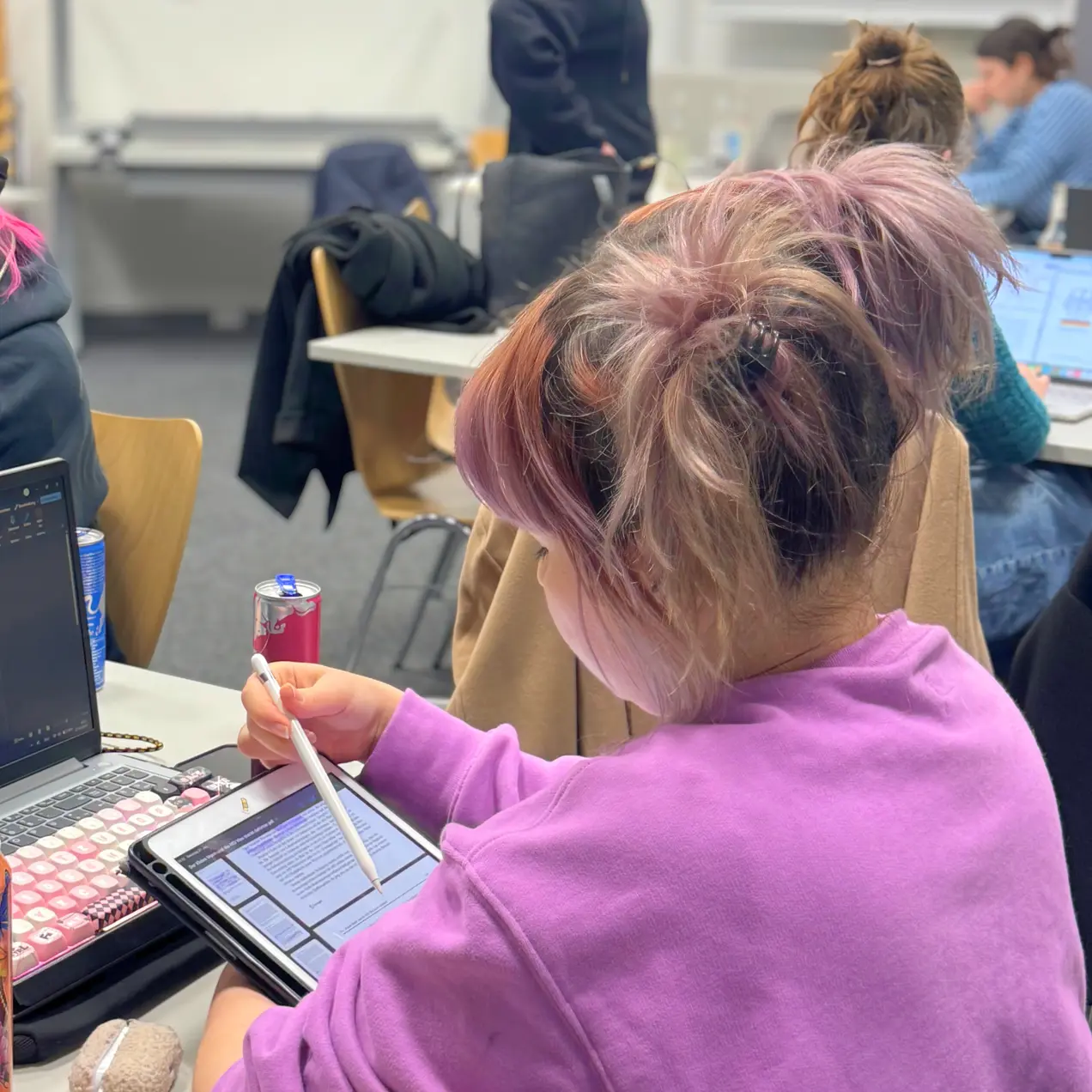 Eine Person mit lila Haaren sitzt an einem Tisch und nutzt ein Tablet, während sie mit einem Stift Notizen macht. Der Tisch ist mit technischen Geräten und Schreibmaterialien ausgestattet. Im Hintergrund sind weitere Personen und Möbel sichtbar.