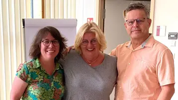 Drei Personen stehen eng beieinander in einem hellen Raum. Die linke Person trägt ein bunt gemustertes Hemd, die mittlere hat ein grau-kleid an, und die rechte Person trägt ein helles Shirt. Alle drei lächeln in die Kamera.