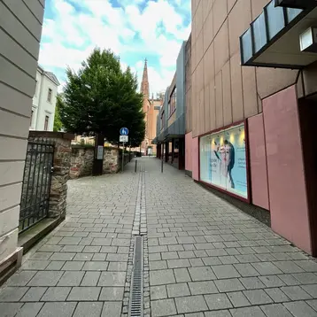 Eine gepflasterte Straße verläuft zwischen Gebäuden. Auf der linken Seite befindet sich eine Mauer und ein Baum, während ein Werbeplakat auf der rechten Seite sichtbar ist. Im Hintergrund steht ein Kirchturm, der hoch in den Himmel ragt, unter einem bewölkten Himmel.