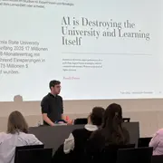 Ein Referent steht vor einem Publikum und spricht über die Auswirkungen von KI auf Universitäten und das Lernen. Auf einer großen Leinwand ist der Text "AI is Destroying the University and Learning Itself" zu sehen, zusammen mit einem Hinweis auf ein Beispiel der California State University.