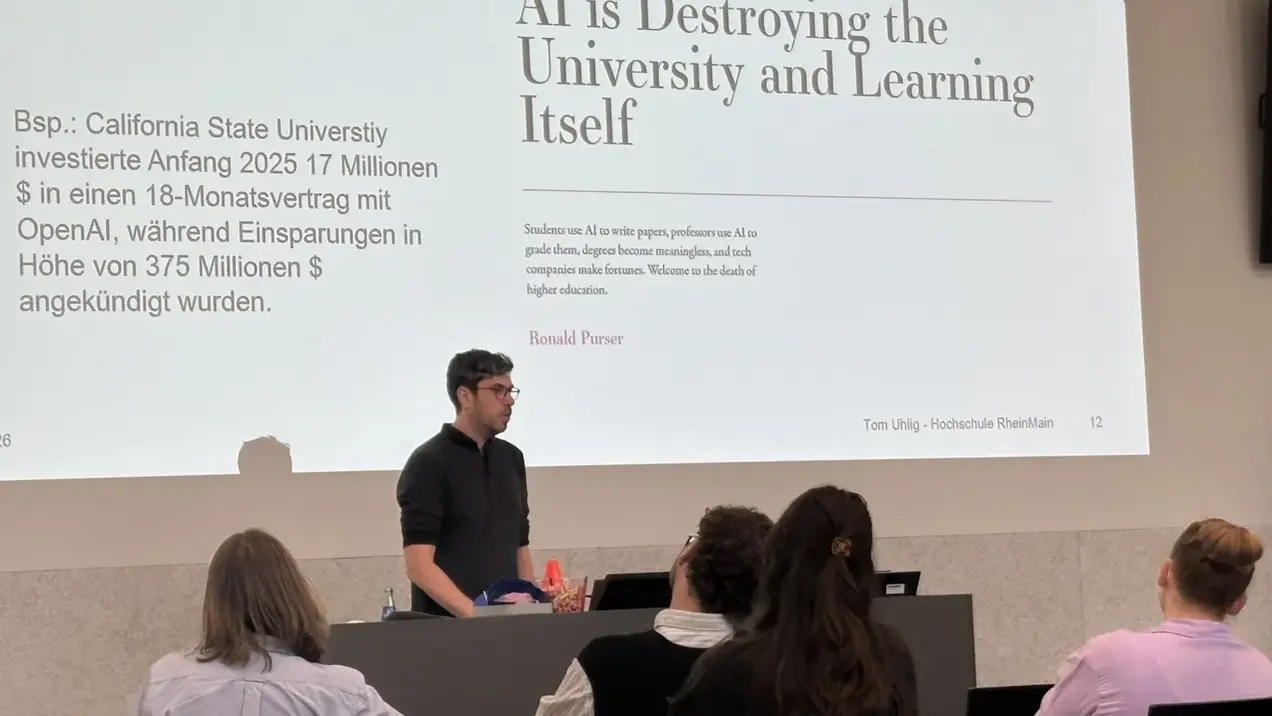 Ein Referent steht vor einem Publikum und spricht über die Auswirkungen von KI auf Universitäten und das Lernen. Auf einer großen Leinwand ist der Text "AI is Destroying the University and Learning Itself" zu sehen, zusammen mit einem Hinweis auf ein Beispiel der California State University.