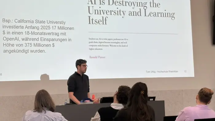 Ein Referent steht vor einem Publikum und spricht über die Auswirkungen von KI auf Universitäten und das Lernen. Auf einer großen Leinwand ist der Text "AI is Destroying the University and Learning Itself" zu sehen, zusammen mit einem Hinweis auf ein Beispiel der California State University.