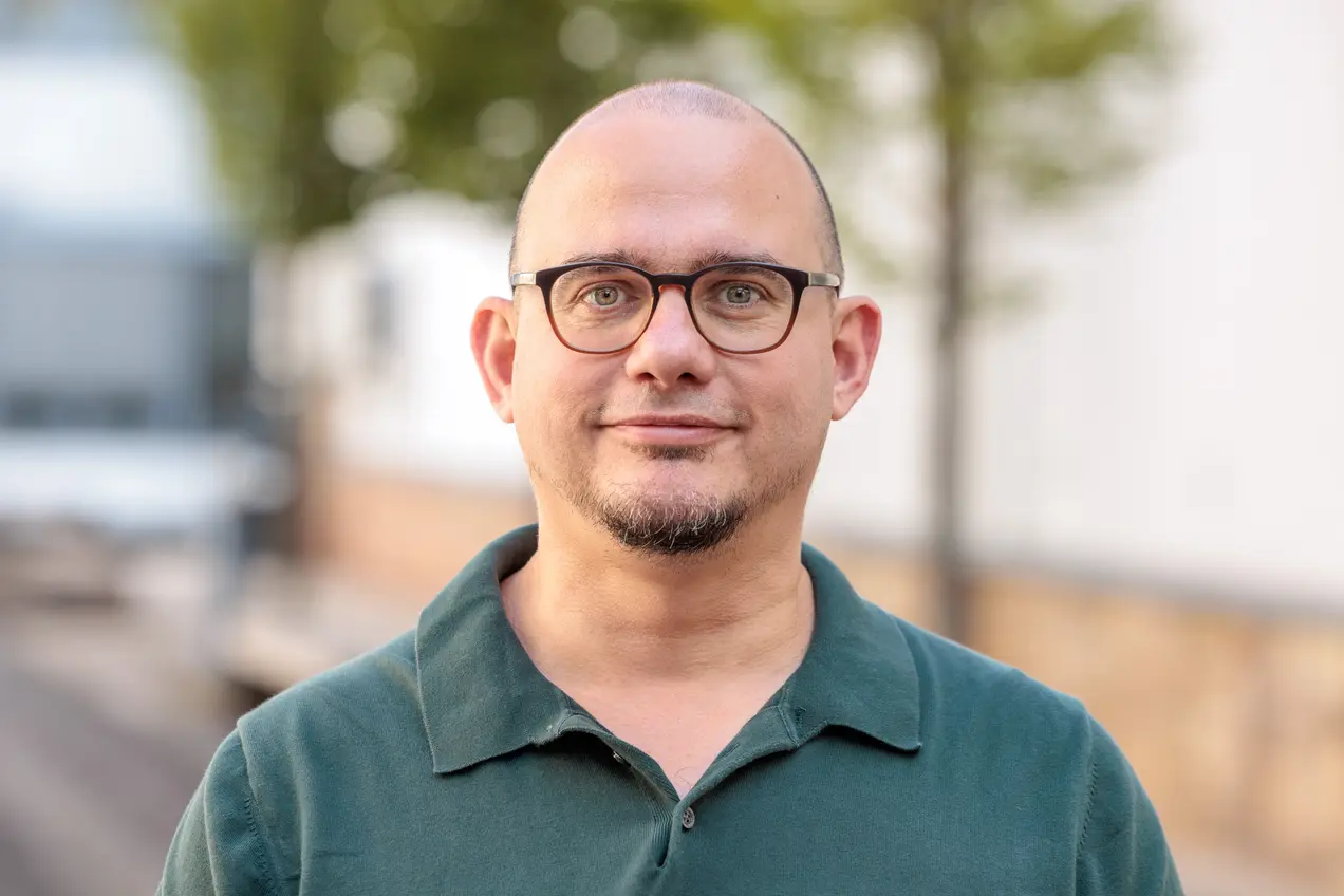 Ein lächelnder Mann mit Glatze und Brille trägt ein dunkelgrünes Polohemd. Er steht im Freien vor einem verschwommenen Hintergrund mit Bäumen und einem Gebäude.