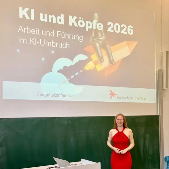 Eine weibliche Person in einem roten Kleid steht vor einer Präsentation mit dem Titel „KI und Köpfe 2026“. Im Hintergrund sind Grafiken zu sehen, die futuristische Themen ansprechen. Der Raum sieht aus wie ein Seminarraum mit einer Tafel.