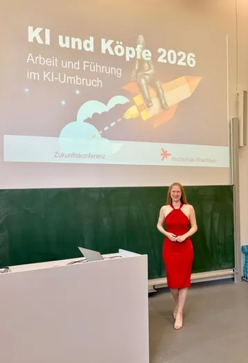 Eine weibliche Person in einem roten Kleid steht vor einer Präsentation mit dem Titel „KI und Köpfe 2026“. Im Hintergrund sind Grafiken zu sehen, die futuristische Themen ansprechen. Der Raum sieht aus wie ein Seminarraum mit einer Tafel.
