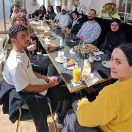 Eine Gruppe von jungen Menschen sitzt an einem Tisch in einem modernen Café. Sie genießen Getränke und Snacks. Die Atmosphäre ist entspannt und freundlich, mit Licht von Fenstern, die eine angenehme Stimmung schaffen.