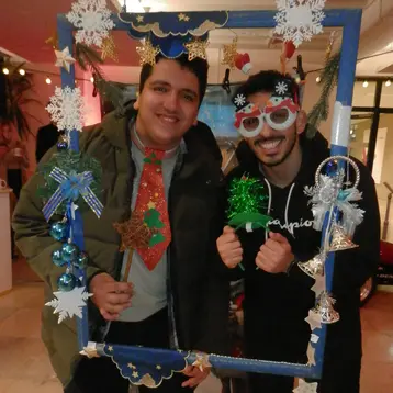 Zwei Männer stehen lächelnd in einem festlich dekorierten Bilderrahmen. Der linke Mann trägt eine Weihnachtskrawatte, während der rechte bunte Brille und eine grüne Weihnachtsdekoration hält. Der Hintergrund zeigt eine festliche Umgebung mit Lichtern und Dekorationen.