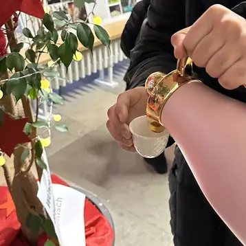 Eine Person gießt aus einer rosa Thermoskanne in einen kleinen weißen Becher. Im Hintergrund sind eine Pflanze und dekorative Elemente zu sehen.