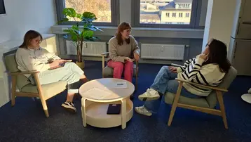 Drei Frauen sitzen in einem hellen Raum zusammen auf Stühlen um einen runden Tisch. Eine Frau redet, während die anderen beiden aufmerksam zuhören. Im Hintergrund ist ein großes Fenster mit Tageslicht und eine Pflanze sichtbar.