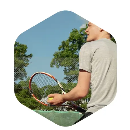 Ein junger Mann hält einen Tennisschläger und schaut auf einen Tennisball, den er in der anderen Hand hält. Im Hintergrund sind Bäume und ein Tennisnetz sichtbar. Der Himmel ist klar, was auf schönes Wetter hinweist.