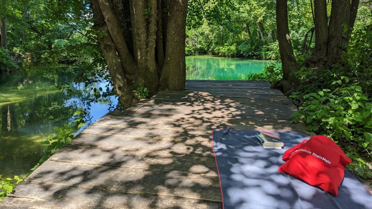 Es ist ein Holzsteg zu sehen, auf dem ein Handtuch mit Buch und HSRM-Tasche liegt. Ein türkiser Fluss im Hintergrund schimmert in der Sonne