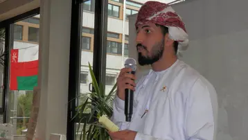 Ein Mann im traditionellen Outfit aus Oman hält ein Mikrofon und spricht. Im Hintergrund sind Fenster mit Blick auf Pflanzen und eine Fahne des Oman zu sehen. Der Raum wirkt festlich und einladend.