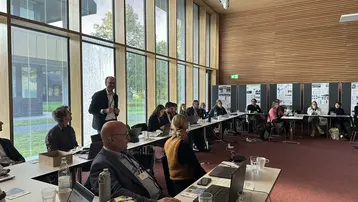 Eine Gruppe von Tagungsteilnehmenden sitzt in einem Konferenzraum, während ein Mann im Vordergrund spricht. Die Wände sind mit Fenstern und Präsentationsmaterialien versehen. Es wird über die Arbeit der UNESCO Lehrstühle in Deutschland gesprochen.