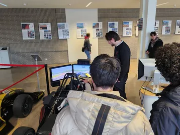 Eine Gruppe von Menschen steht vor einem Simulator für ein Rennauto. Ein Mann erklärt etwas am Bildschirm, während andere aufmerksam zuschauen. Das Setting ist ein modernes, hell erleuchtetes Veranstaltungsgebäude.