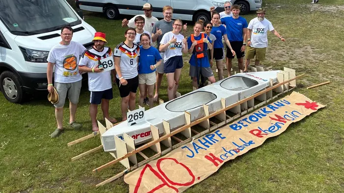 Eine Gruppe von zehn Personen steht vor einem Bootsrahmen. Sie tragen sportliche T-Shirts und lächeln, während sie den Slogan „40 Jahre Betonkahn“ auf einem Banner in der Mitte präsentieren. Im Hintergrund sind mehrere Transportfahrzeuge zu sehen.