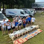 Eine Gruppe von zehn Personen steht vor einem Bootsrahmen. Sie tragen sportliche T-Shirts und lächeln, während sie den Slogan „40 Jahre Betonkahn“ auf einem Banner in der Mitte präsentieren. Im Hintergrund sind mehrere Transportfahrzeuge zu sehen.