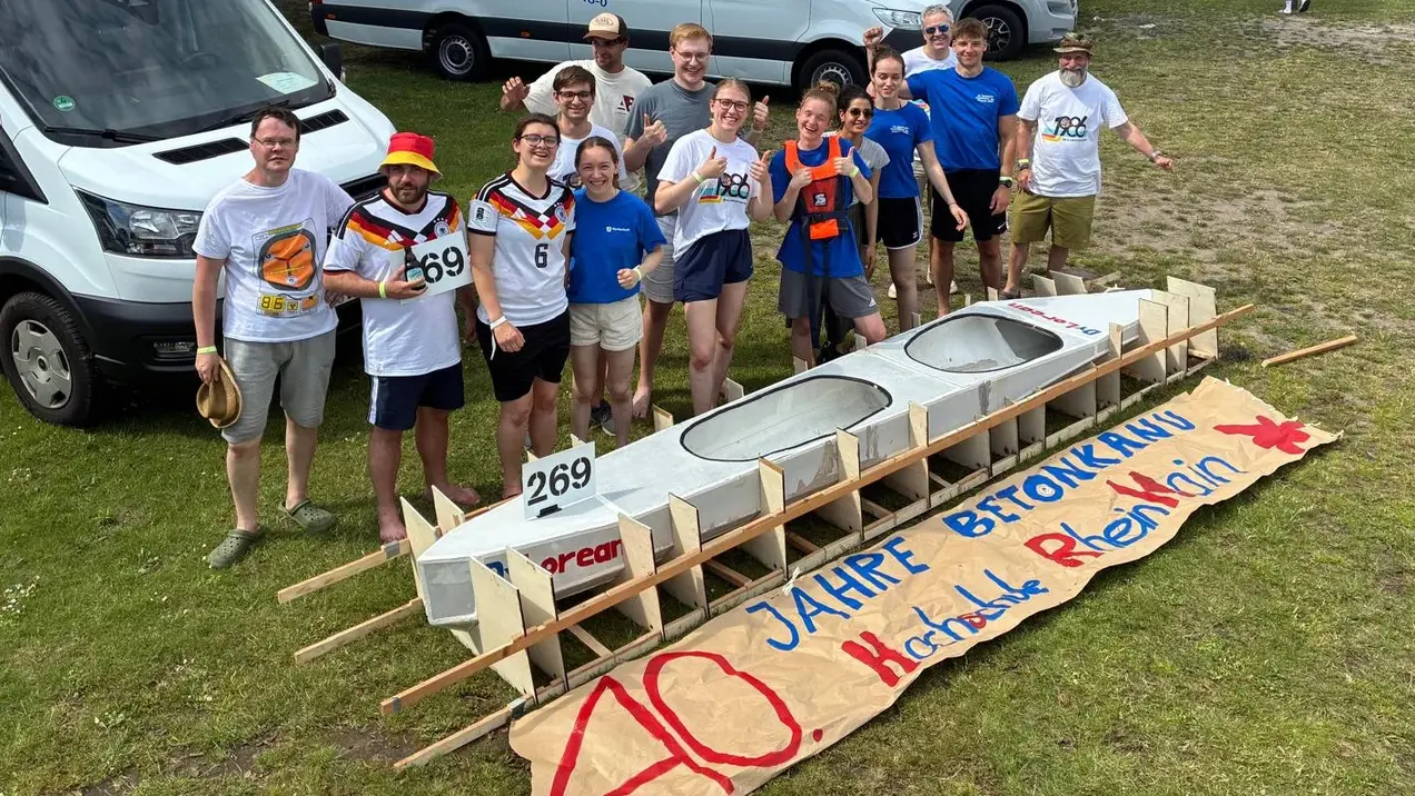 Eine Gruppe von zehn Personen steht vor einem Bootsrahmen. Sie tragen sportliche T-Shirts und lächeln, während sie den Slogan „40 Jahre Betonkahn“ auf einem Banner in der Mitte präsentieren. Im Hintergrund sind mehrere Transportfahrzeuge zu sehen.