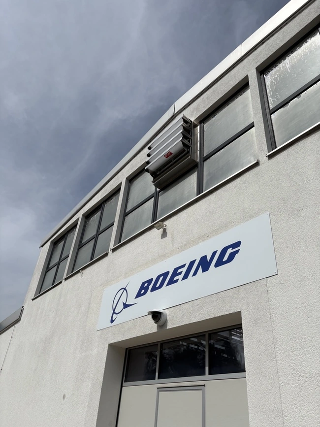 Das Bild zeigt die Fassade eines Gebäudes mit einem großen, weißen Schild, auf dem "BOEING" in blauen Buchstaben steht. Über dem Schild sind mehrere Fenster sichtbar, und an der Wand befindet sich ein Klimagerät. Der Himmel ist teilweise bewölkt.