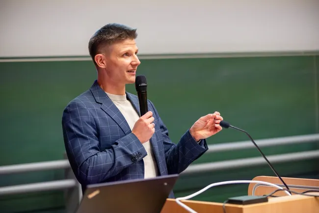 Ein Mann steht an einem Rednerpult und hält ein Mikrofon in der Hand. Er trägt einen blauen Blazer und erläutert offensichtlich ein Thema. Im Hintergrund ist eine grüne Tafel sichtbar. Der Raum scheint eine akademische Umgebung zu sein.