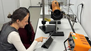 Zwei Frauen sitzen an einem Tisch in einem Labor. Eine der Frauen zeigt mit dem Finger auf ein Tablet. Im Hintergrund sind ein großer Zylinder und ein orangefarbenes Gerät zu sehen. Die Umgebung wirkt technisch und wissenschaftlich.