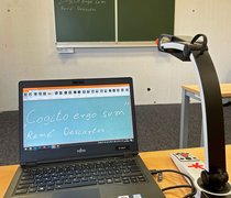 Eine Tafelbildkamera nimmt Text von einer Tafel auf, dieser ist auf dem danebenstehenden Laptop stark vergrößert zu lesen