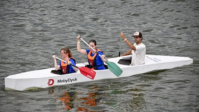 Drei Personen in einem weißen Boot paddeln auf einem Gewässer. Zwei Frauen sitzen vorne und halten Paddel, während ein Mann hinten steht. Alle tragen Schwimmwesten und sind konzentriert auf das Paddeln.