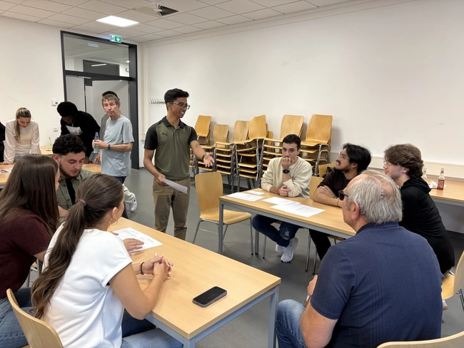 Eine Gruppe junger Menschen sitzt an Tischen in einem Raum. Eine Person steht und erklärt etwas, während die anderen aufmerksam zuhören und Notizen machen. Der Raum wirkt informell und einladend. Im Hintergrund stehen Holzstühle gestapelt.