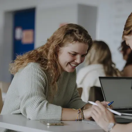 Man sieht zwei Studierende im Vordergrund, die gemeinsam an einem Projekt arbeiten und lächeln. Im Hintergrund sieht man weitere Studierende, die sich konzentrieren.