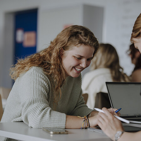 Man sieht zwei Studierende im Vordergrund, die gemeinsam an einem Projekt arbeiten und lächeln. Im Hintergrund sieht man weitere Studierende, die sich konzentrieren.