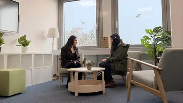Zwei Frauen sitzen in einem modernen, hellen Raum mit großen Fenstern. Sie sprechen miteinander, während ein kleiner Tisch zwischen ihnen steht. Pflanzen und ein Fernseher sind im Hintergrund sichtbar, was eine entspannte Atmosphäre schafft.