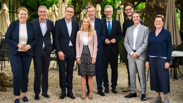 Eine Gruppe von neun Personen posiert im Freien. Sie stehen in einer Reihe vor einem Gartenlokal, umgeben von Stühlen und Schirmen. Die Anwesenden tragen formelle Kleidung und lächeln in die Kamera, während sie eine freundliche Atmosphäre ausstrahlen.