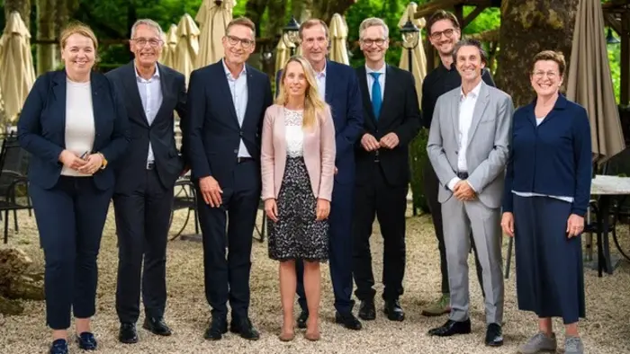 Eine Gruppe von neun Personen posiert im Freien. Sie stehen in einer Reihe vor einem Gartenlokal, umgeben von Stühlen und Schirmen. Die Anwesenden tragen formelle Kleidung und lächeln in die Kamera, während sie eine freundliche Atmosphäre ausstrahlen.