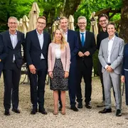 Eine Gruppe von neun Personen posiert im Freien. Sie stehen in einer Reihe vor einem Gartenlokal, umgeben von Stühlen und Schirmen. Die Anwesenden tragen formelle Kleidung und lächeln in die Kamera, während sie eine freundliche Atmosphäre ausstrahlen.