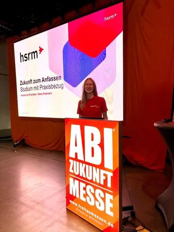 Eine Frau steht an einem Informationsstand mit einem Banner, auf dem "Zukunft zum Anfassen" und "Studium mit Praxisbezug" steht. Im Hintergrund ist eine große Leinwand mit dem Logo der hsrm und weiteren Informationen zu sehen. Der Stand ist Teil einer Messe zur Studien- und Berufsorientierung.