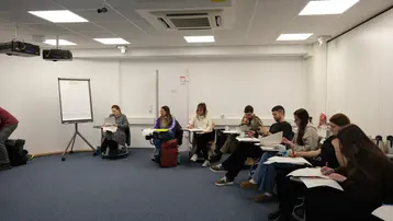 Eine Gruppe von Personen sitzt in einem Schulungsraum und macht Notizen. Einige Teilnehmer schauen nach vorne, während andere auf ihre Notizblätter geschrieben. Im Hintergrund steht ein Flipchart. Der Raum hat einen blauen Teppich und eine helle Atmosphäre.