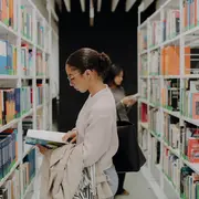 Eine Frau steht in einer Bibliothek zwischen Regalen voller Bücher. Sie trägt eine Brille und hält ein geöffnetes Buch in der Hand, während sie konzentriert liest. Im Hintergrund ist eine weitere Person zu sehen, die ebenfalls in den Regalen stöbert.