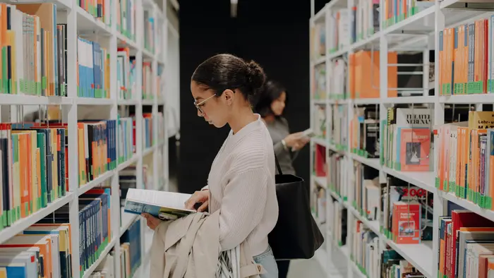 Eine Frau steht in einer Bibliothek zwischen Regalen voller Bücher. Sie trägt eine Brille und hält ein geöffnetes Buch in der Hand, während sie konzentriert liest. Im Hintergrund ist eine weitere Person zu sehen, die ebenfalls in den Regalen stöbert.